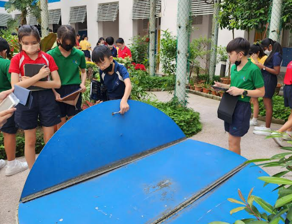 Children wearing masks examine a large blue object outdoors near a school garden, holding notebooks.